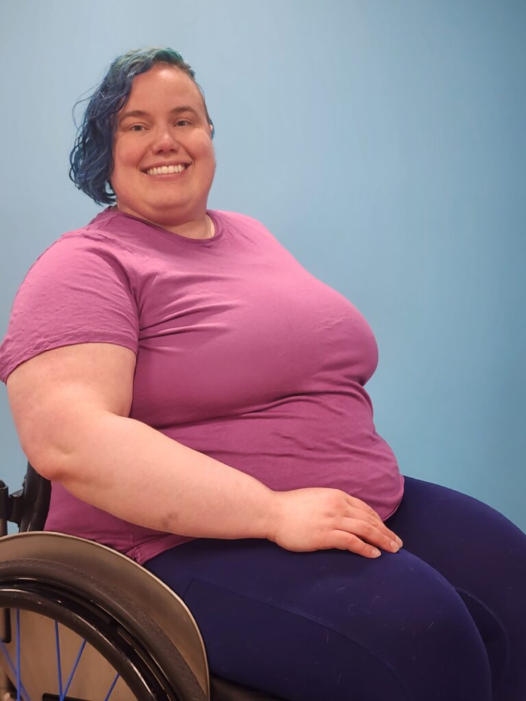 Paulina is a white non-binary person in their 30s with chin-length blue hair. They are seated in their manual wheelchair and wearing a purple shirt and dark blue leggings. 