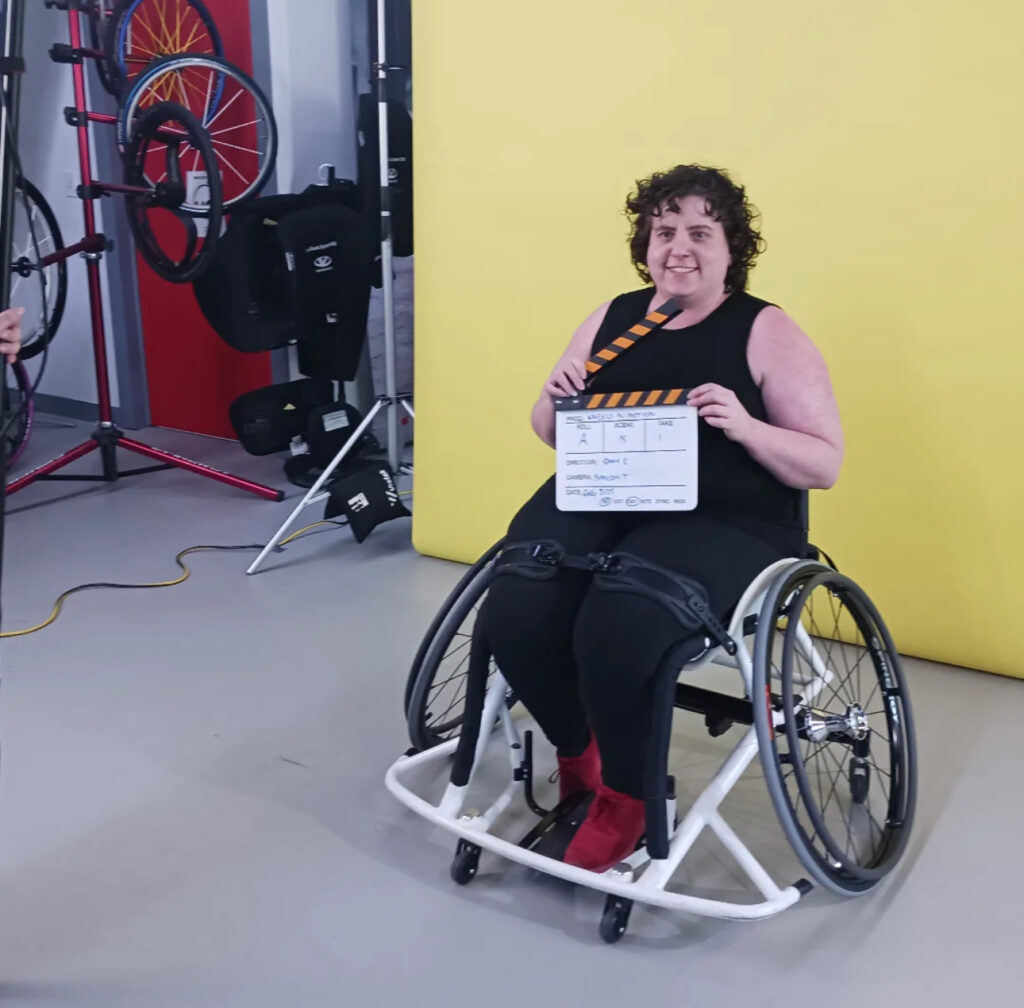 Shay sitting in a white sports wheelchair holding a video clapboard. They are against a yellow backdrop with various wheelchair parts visible in the background.