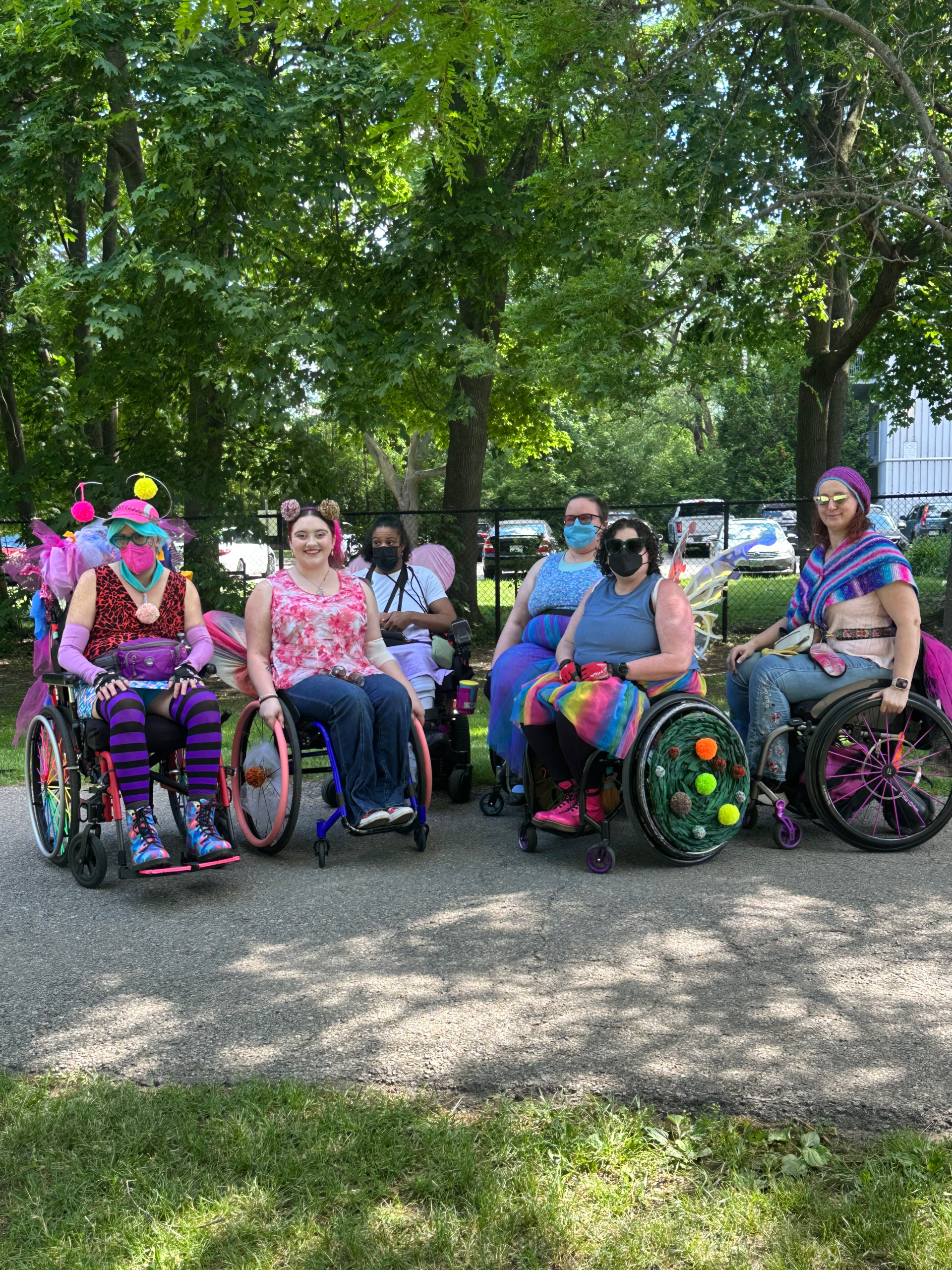 A group shot of participants in Wheels on Parade. A group of six wheelchair users with brightly coloured chairs gather in a park.